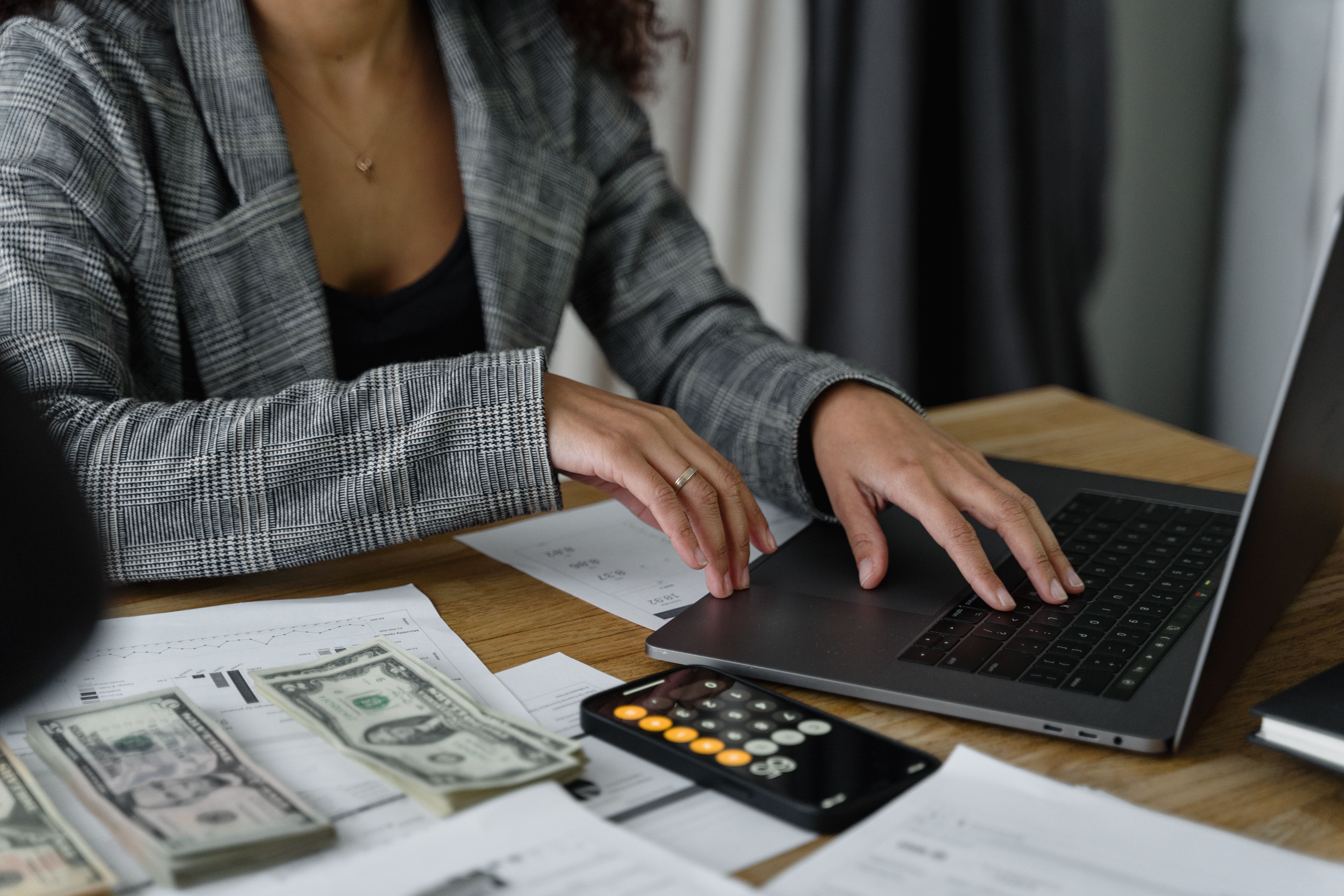 Finance professional processing payroll on a laptop with US dollars, a calculator, and financial documents on the desk